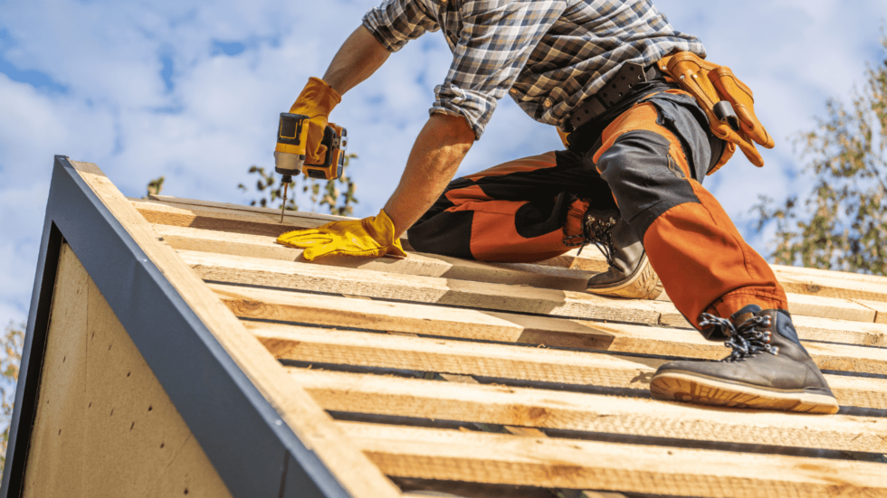 A roofer wearing sturdy gloves uses a power drill to construct the frame of a roof 980x551