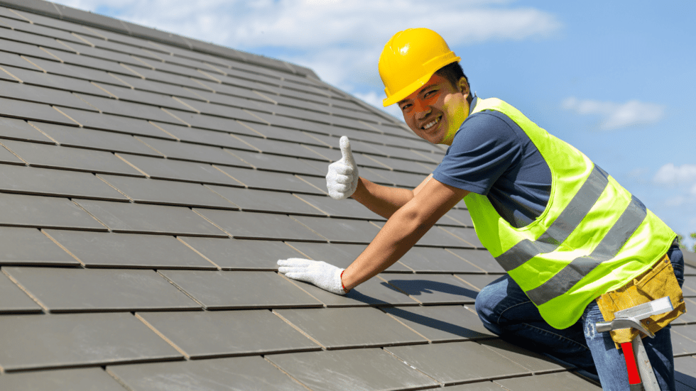 A roofer on a tile roof and wearing gloves and a hard hat gives a thumbs up 980x551