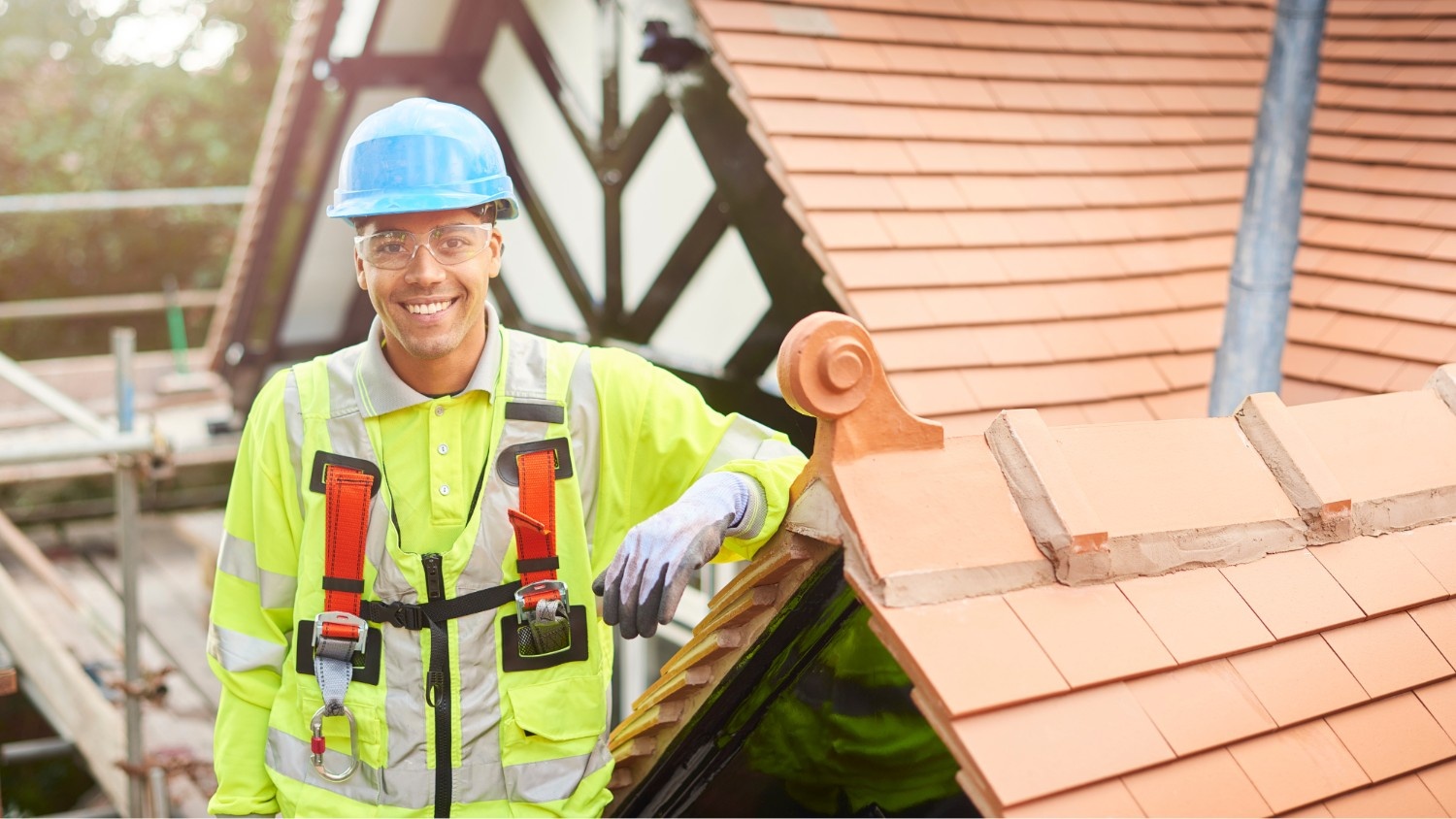 Construction worker in safety gear and hard hat stands on a roof next to terracotta tiles, smiling at the camera. Scaffolding and another building are in the background.