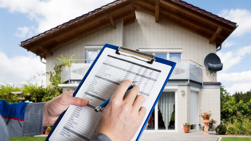 A roofer holds a clipboard and pen while looking at the roof of a house.