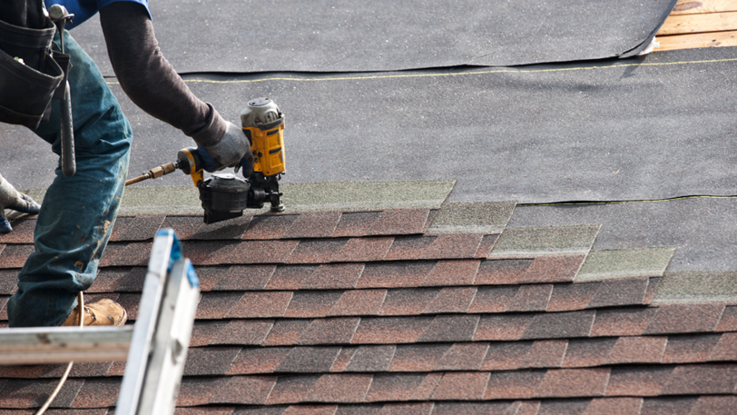 A roofer uses a nail gun to install asphalt shingles on a residential roof.