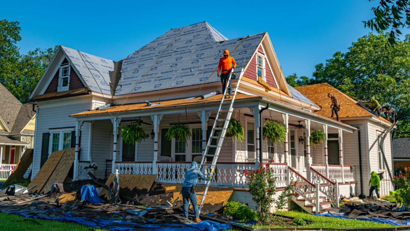 A large home with multiple roofers working on ladders to remove and replace the roofing.